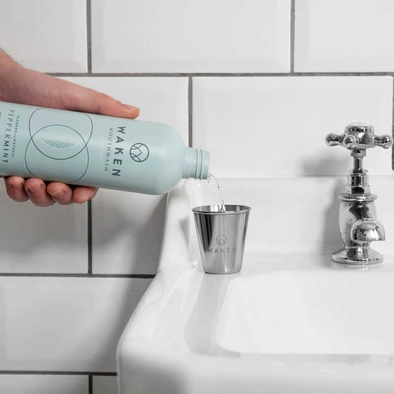 Person pouring a bottle of Waken eco-friendly mouthwash into a metal cup in a white tiled bathroom.