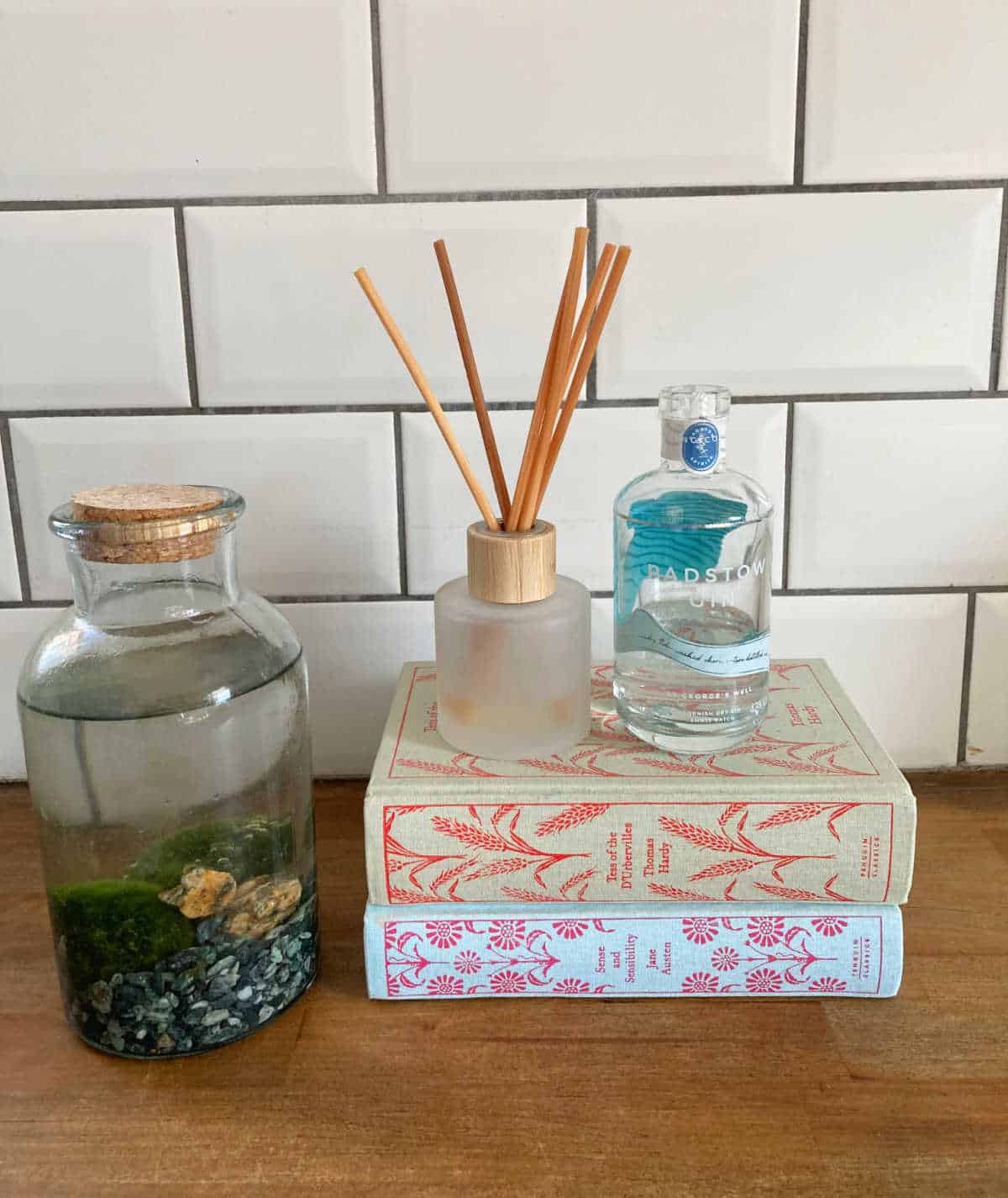 Reed diffuser sitting on books, next to a mini bottle of Padstow gin and a glass bottle of moss balls.
