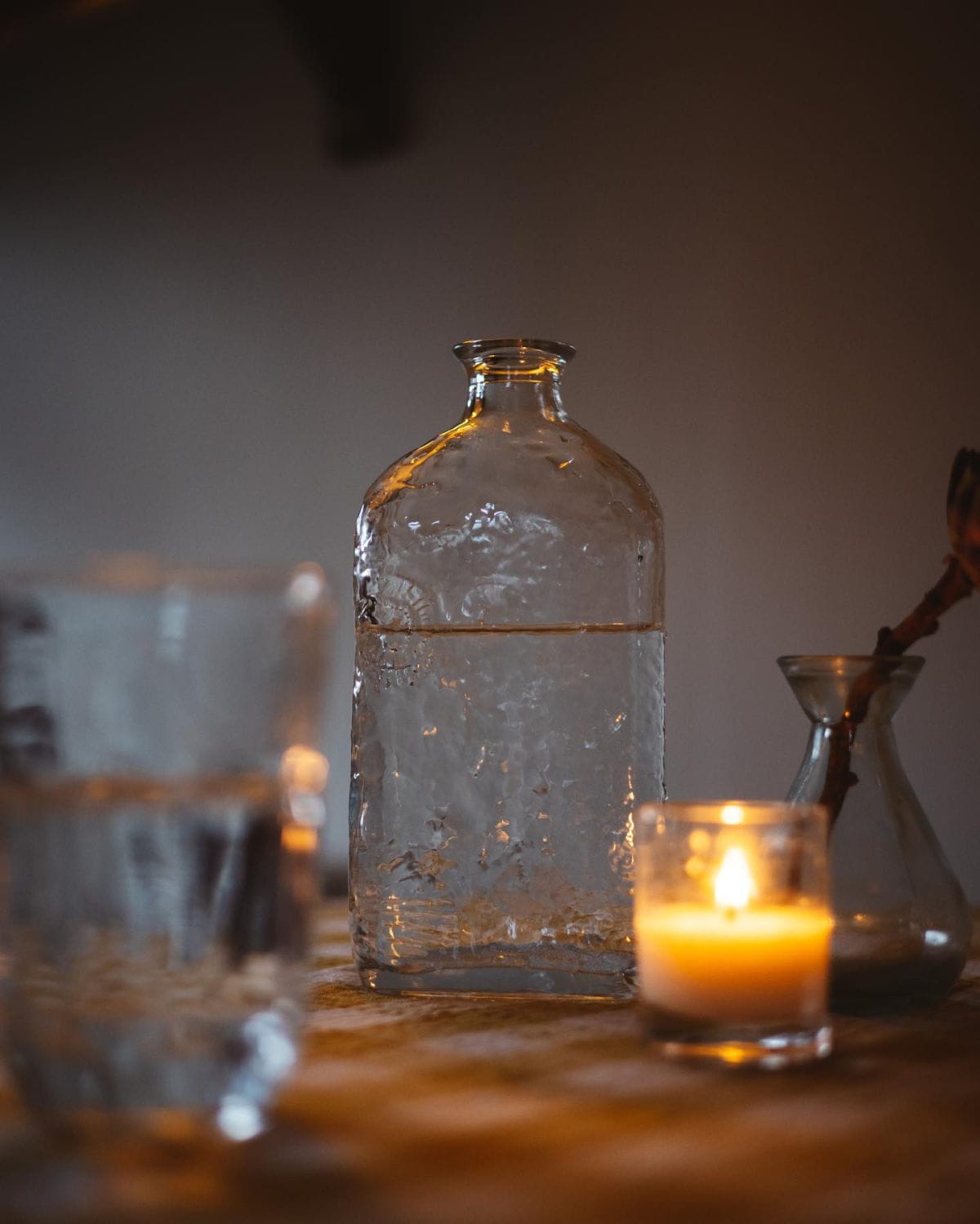 Old spirit bottle reused as a water bottle, on a table with glasses and candles.