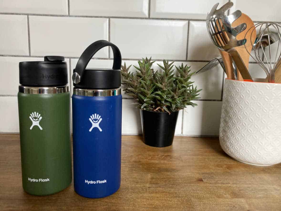 A green Hydro Flask and a navy blue Hydro Flask sitting on a wooden kitchen worktop next to a plant and utensil holder.